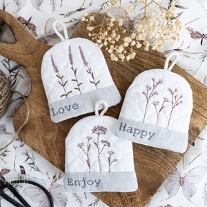 以下が含まれることがあります： 花柄の刺繍が施された白いキルティングの鍋つかみが3つあり、「Love」、「Happy」、「Enjoy」の文字が書かれています。