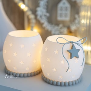 May include: Two white ceramic candle holders with star cutouts, illuminated from within. One has a blue star charm hanging from a string. Both have a grey beaded base. The background includes fairy lights and a decorative wreath.