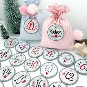 Könnte beinhalten: Ein Set mit 22 grauen Filz-Adventskalender-Anhängern mit rot-weiß gestreiftem Stoff und roter Stickerei. Die Anhänger sind von 1 bis 22 nummeriert und beinhalten Wörter wie "Elf", "Sieben" und "Neun".