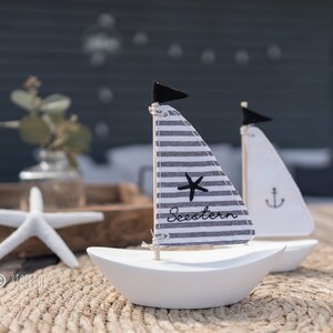 Könnte beinhalten: Zwei weiße Miniatur-Segelschiffe mit Stoffsegeln. Ein Segel ist gestreift und sagt "Seestern" und das andere ist weiß mit einem Anker. Die Segelschiffe stehen auf einem gewebten Teppich.