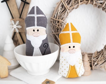 May include: Two handmade felt Saint Nicholas figures, one in gray with a white beard and a white cross on the hat, and the other in yellow with a white beard and a white cross on the hat, sitting in a white bowl.