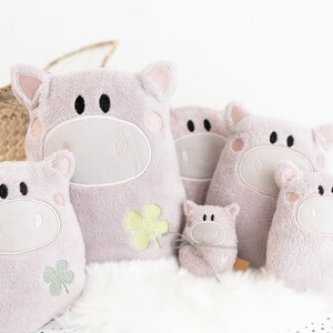 Puede incluir: Cinco juguetes de peluche de cerdo rosa de diferentes tama&ntilde;os, cada uno con ojos bordados y un dise&ntilde;o de tr&eacute;bol. El cerdo m&aacute;s grande est&aacute; sentado derecho, mientras que los dem&aacute;s est&aacute;n dispuestos a su alrededor.