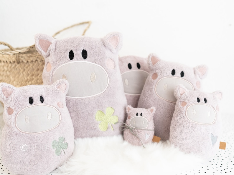 Puede incluir: Cinco juguetes de peluche de cerdo rosa de diferentes tama&ntilde;os, cada uno con ojos bordados y un dise&ntilde;o de tr&eacute;bol. El cerdo m&aacute;s grande est&aacute; sentado derecho, mientras que los dem&aacute;s est&aacute;n dispuestos a su alrededor.