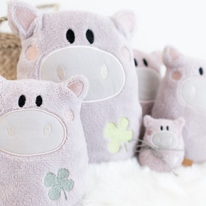 Puede incluir: Cuatro juguetes de peluche en forma de cerdo de diferentes tama&ntilde;os, todos rosas con caras bordadas y detalles de tr&eacute;bol. El cerdo m&aacute;s grande est&aacute; sentado derecho, mientras que los dem&aacute;s est&aacute;n dispuestos en fila delante de &eacute;l.