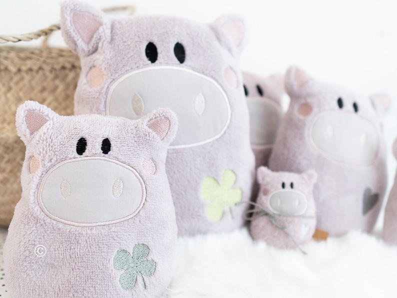 Puede incluir: Cuatro juguetes de peluche en forma de cerdo de diferentes tama&ntilde;os, todos rosas con caras bordadas y detalles de tr&eacute;bol. El cerdo m&aacute;s grande est&aacute; sentado derecho, mientras que los dem&aacute;s est&aacute;n dispuestos en fila delante de &eacute;l.