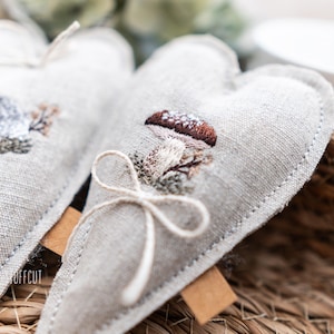 May include: Two heart-shaped, light grey linen sachets with embroidered woodland designs. One features a hedgehog, the other a mushroom. Each sachet has a twine bow and a small brown tag. The sachets are in a woven basket.