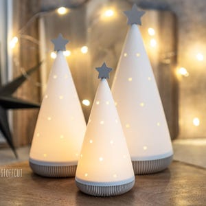 May include: Three white ceramic Christmas tree-shaped lights with grey star toppers and dotted patterns. The lights have a grey base with a striped design. The trees are lit, creating a warm glow, and are arranged on a wooden tray.