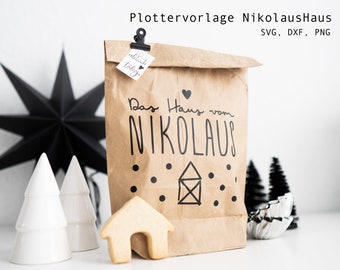 Op de afbeelding: Een bruine papieren zak met een zwart-wit ontwerp met de tekst "Das Haus vom Nikolaus" met een klein huis en stippen. De zak is vastgemaakt met een zwarte clip en een klein label met de tekst "celebrate today".