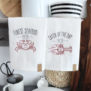 Könnte beinhalten: Zwei weiße Küchentücher mit roter Stickerei. Ein Handtuch sagt "Finest Seafood Fresh" mit einem Krabben-Design. Das andere Handtuch sagt "Catch of the Day Fresh" mit einem Hummer-Design.