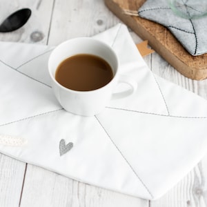 May include: White fabric coaster with a stitched heart design and a cup of coffee on top. A starfish is on the table next to the coaster.