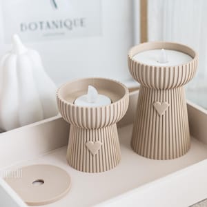 以下が含まれることがあります： ハートのデザインのベージュ色のキャンドルホルダー2つ。それぞれ白いLEDキャンドルが入っており、お揃いのトレイに置かれています。キャンドルホルダーはリブのデザインです。「BOTANIQUE」と書かれた白いカボチャと額入りのプリントが背景にあります。