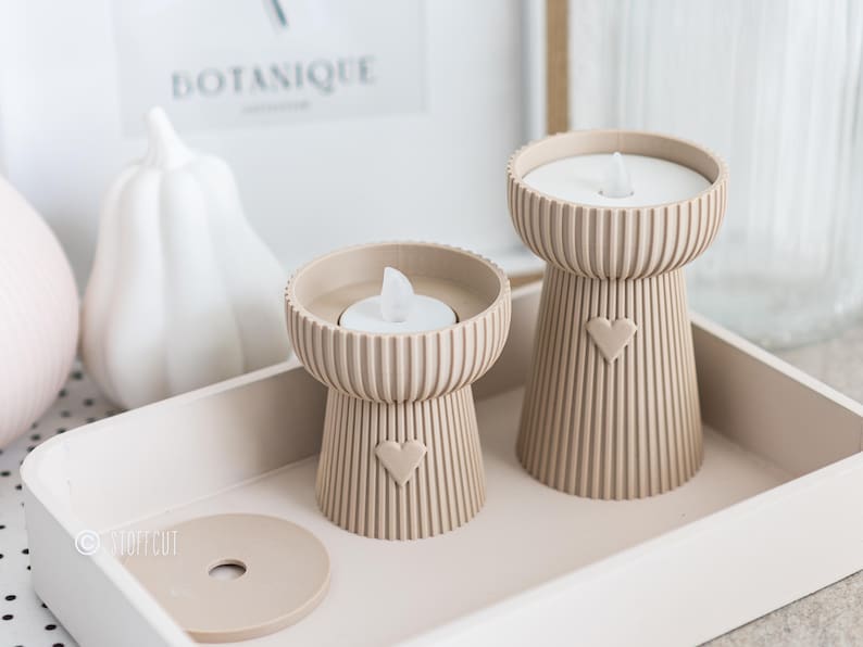 May include: Two beige candle holders with a heart design, each holding a white LED candle, sit on a matching tray. The candle holders have a ribbed design. A white pumpkin and a framed print that says "BOTANIQUE" are in the background.