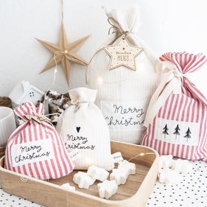Può includere: Quattro sacchetti regalo in tessuto bianco con sacchetti regalo a righe rosse e bianche. I sacchetti sono decorati con il testo "Merry Christmas" e "Winter Hugs". I sacchetti sono su un vassoio di legno con bombe da bagno bianche a forma di stella.