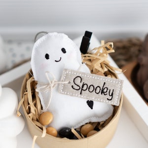 Op de afbeelding: Een wit, spookvormig pluche speelgoed met een glimlachend gezicht en het woord "Spooky" geborduurd op een bruin jute label. Het speelgoed zit in een bruine papieren beker gevuld met gescheurd papier en kleine bruine en zwarte kralen.