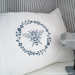 May include: White pillow with a blue embroidered bee in a floral wreath. The pillow is on a striped background.