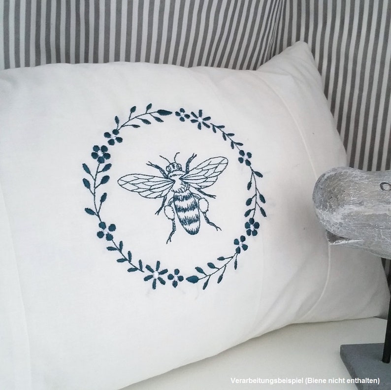 May include: White pillow with a blue embroidered bee in a floral wreath. The pillow is on a striped background.