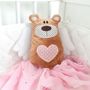 Pu&ograve; includere: Un orsacchiotto di peluche marrone con un cuore rosa sul petto e ali bianche. L'orso &egrave; seduto su un tessuto di tulle rosa con stelle argentate.