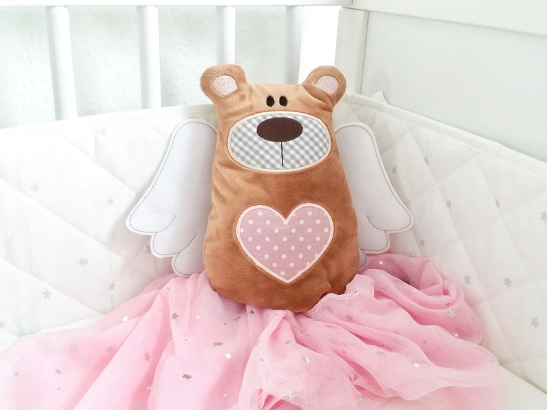 Pu&ograve; includere: Un orsacchiotto di peluche marrone con un cuore rosa sul petto e ali bianche. L'orso &egrave; seduto su un tessuto di tulle rosa con stelle argentate.