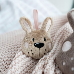 May include: A brown plush bunny toy with a pink ribbon loop. The bunny has black eyes, a black nose, and a pink nose pad. The bunny is lying on a pink knitted blanket.