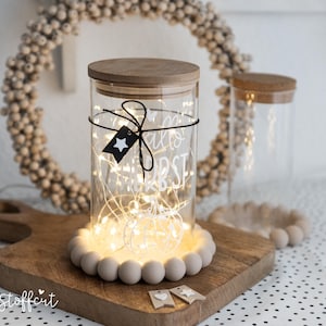 Könnte beinhalten: Ein zylindrisches Glasgefäß mit Holzdeckel, gefüllt mit warmweißer Lichterkette. Das Glas ist mit dem weißen Schriftzug "hallo herbst" und einem kleinen schwarzen Anhänger mit weißem Stern versehen und steht auf einem Holzbrett mit Holzkugeln.