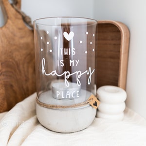 Può includere: Un portacandele in vetro con una base in cemento. Il vetro è decorato con un cuore bianco e il testo "This is my happy place". Il portacandele è su una superficie bianca.