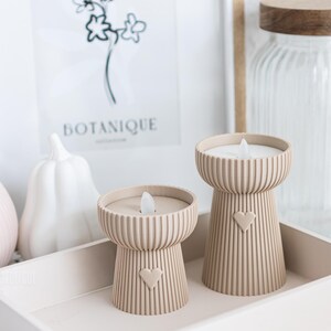 May include: Two beige candle holders with a heart design, each with a faux flame. The candle holders are on a tray with other decorative items, including a white pumpkin and a framed botanical print. The candle holders are a decorative home accent.