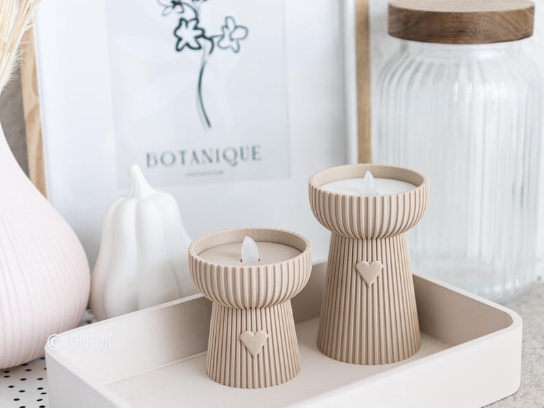 May include: Two beige candle holders with a heart design, each with a faux flame. The candle holders are on a tray with other decorative items, including a white pumpkin and a framed botanical print. The candle holders are a decorative home accent.