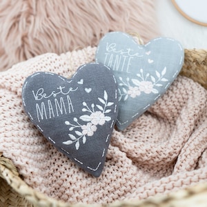 May include: Two gray heart-shaped pillows with embroidered floral designs and the text "Beste Mama" and "Beste Tante".
