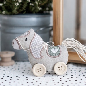 Puede incluir: Un caballo de juguete de tela gris y blanco con cola blanca y ruedas. El caballo tiene un parche en forma de corazón en la espalda y una brida blanca.