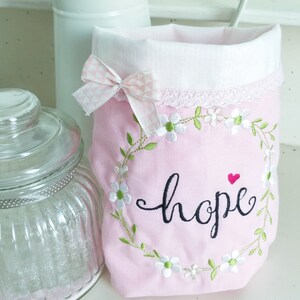 May include: Pink fabric bag with a white lace trim and a white bow. The bag is embroidered with a wreath of green leaves and white flowers, and the word "hope" in black cursive lettering. A small red heart is above the word "hope".