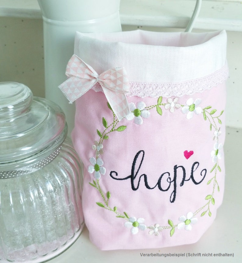 May include: Pink fabric bag with a white lace trim and a white bow. The bag is embroidered with a wreath of green leaves and white flowers, and the word "hope" in black cursive lettering. A small red heart is above the word "hope".
