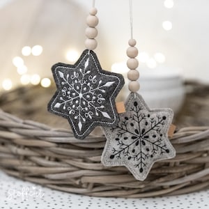 Könnte beinhalten: Zwei Filz-Stern-Ornamente mit Schneeflocken-Stickerei. Eines ist dunkelgrau mit weißen Nähten, das andere hellgrau mit schwarzen Nähten. Jeder Stern hängt an einer Schnur aus Holzperlen. Die Ornamente werden in einem Weidenkorb präsentiert.