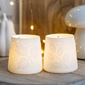 Op de afbeelding: Twee witte keramische kandelaars met bladeren en kleine stippen. De kandelaars worden van binnenuit verlicht, waardoor een warme gloed ontstaat. De kandelaars staan op een houten dienblad, met een vage achtergrond van feeërieke lichtjes en decoratieve objecten.