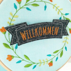 May include: A light blue embroidery hoop with a gray felt banner that says "WILLKOMMEN" in orange embroidery thread. The hoop is decorated with a wreath of green and yellow leaves and orange flowers.