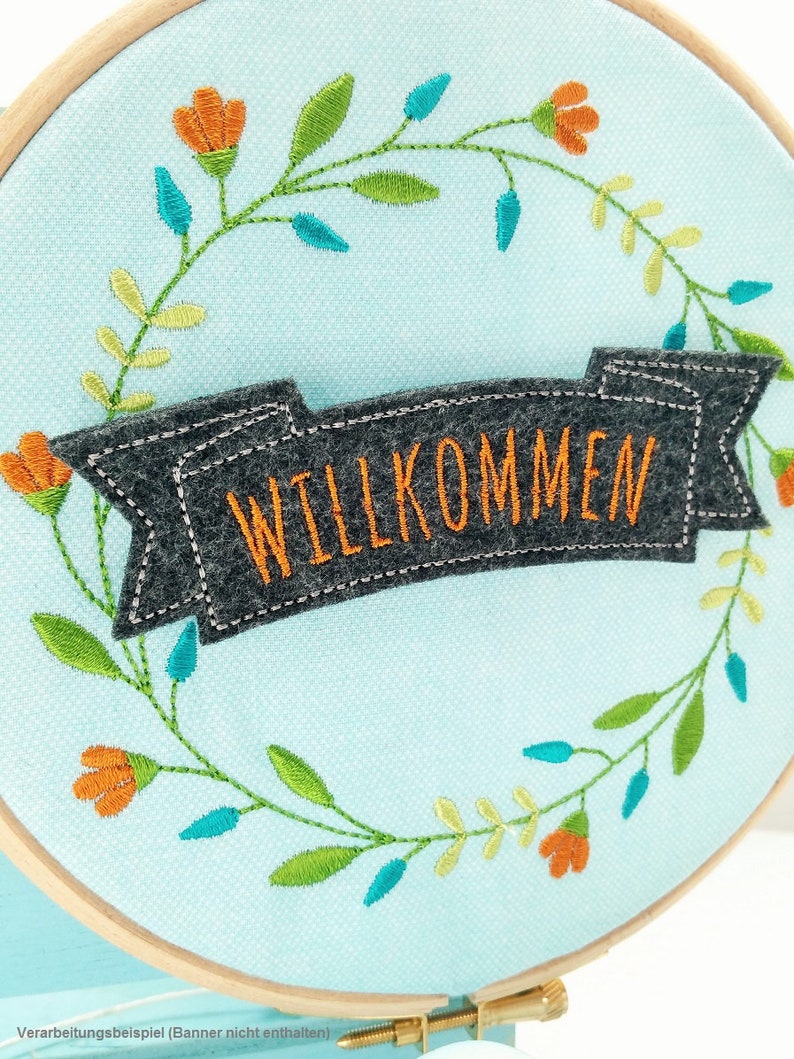 May include: A light blue embroidery hoop with a gray felt banner that says "WILLKOMMEN" in orange embroidery thread. The hoop is decorated with a wreath of green and yellow leaves and orange flowers.