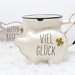 以下が含まれることがあります： 白いぬいぐるみ製のブタのオーナメントで、「VIEL GLÜCK」という文字と、金色の四つ葉のクローバーが刺繍されています。オーナメントは茶色の紐で吊り下げられており、白い表面の上に置かれています。