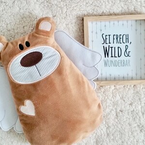 Pu&ograve; includere: Un orso di peluche marrone con ali bianche e un cuore sul petto. L'orso &egrave; seduto su una superficie bianca e soffice. Una cornice di legno con sfondo bianco e testo nero recita "Sei frech, Wild & Wunderbar" con frecce sullo sfondo.