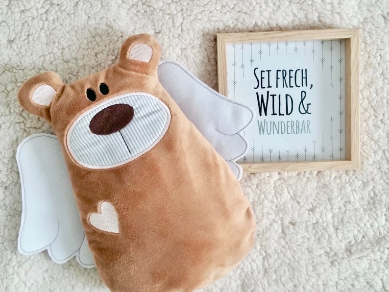 Pu&ograve; includere: Un orso di peluche marrone con ali bianche e un cuore sul petto. L'orso &egrave; seduto su una superficie bianca e soffice. Una cornice di legno con sfondo bianco e testo nero recita "Sei frech, Wild & Wunderbar" con frecce sullo sfondo.