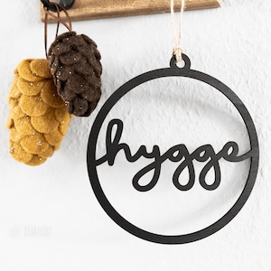 Può includere: Cerchio di legno nero con la parola "hygge" scritta in corsivo. Due pigne di feltro sono appese sopra il cerchio, una è marrone e l'altra è gialla.