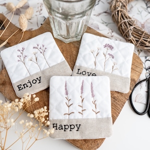 May include: Four white quilted coasters with embroidered floral designs and the words "Enjoy", "Love", "Happy", and "Hope" on a wooden surface.