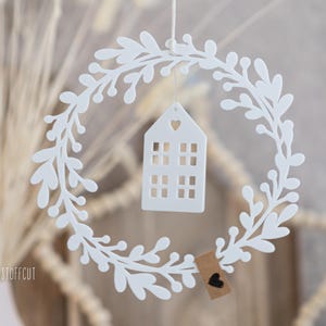 May include: A white, circular wreath with a leafy design, surrounding a small white house-shaped ornament with a heart cutout. A small tag with a black heart hangs below. The wreath is suspended by a thin string.