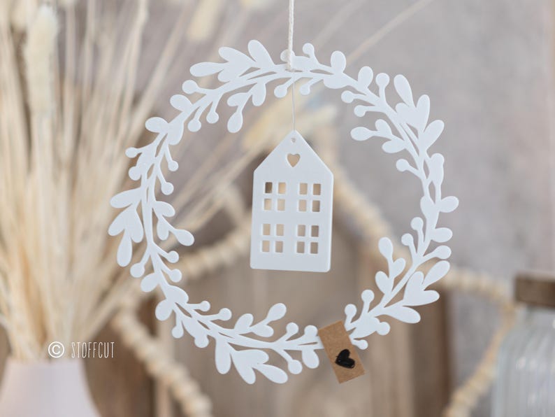May include: A white, circular wreath with a leafy design, surrounding a small white house-shaped ornament with a heart cutout. A small tag with a black heart hangs below. The wreath is suspended by a thin string.