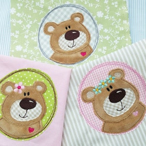Puede incluir: Tres caras de osito de peluche bordadas sobre fondos de tela de diferentes colores. Los osos tienen mejillas rosadas y un corazón rosa en el pecho. Los fondos de tela son verde, rosa y blanco con una raya azul claro.