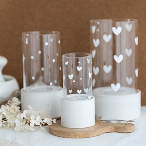 Peut inclure: Trois bougeoirs en céramique blanche avec des inserts en verre pour bougies votives. Les bougies votives en verre sont décorées de points et de cœurs blancs.