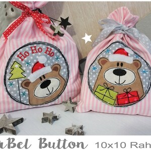 Könnte beinhalten: Zwei rosa-weiß gestreifte Stoffgeschenktüten mit gestickten Designs. Die größere Tasche hat einen grauen Kreis mit einem braunen Bären, der einen Weihnachtsmann-Hut trägt, einen grünen Weihnachtsbaum und den Text "Ho Ho Ho". Die kleinere Tasche hat einen grauen Kreis mit einem braunen Bären, der einen Weihnachtsmann-Hut trägt und zwei Geschenke hält.
