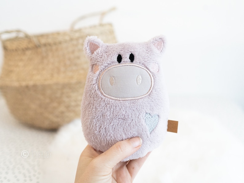 Puede incluir: Un animal de peluche suave y mullido de color lavanda con forma de cerdo con ojos bordados y un coraz&oacute;n en un lado.