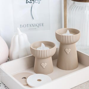 May include: Two beige candle holders with a heart design, each holding a white LED candle, sit on a tray. The holders have a ribbed texture. A white pumpkin and a vase are in the background. The word "BOTANIQUE" is visible in a framed print.