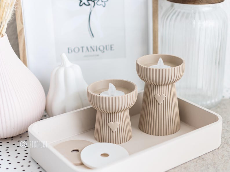 May include: Two beige candle holders with a heart design, each holding a white LED candle, sit on a tray. The holders have a ribbed texture. A white pumpkin and a vase are in the background. The word "BOTANIQUE" is visible in a framed print.