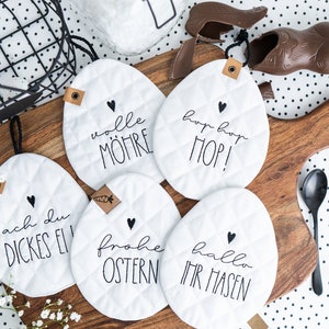 May include: Five white quilted egg-shaped pot holders with black embroidered Easter-themed sayings. The sayings are "ach du dickes Ei", "wolle möhre", "hop hop HOP!", "frohe ostern", and "hallo ihr hasen".