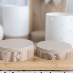 May include: Two beige, round containers with ribbed sides and star details sit on a wooden tray. The containers have a lid and are in front of white ceramic objects. The containers are likely for storage.